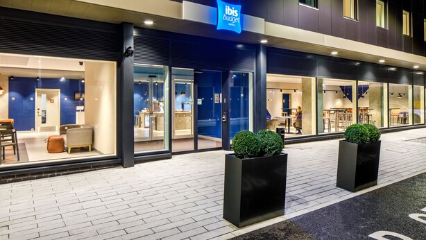 A modern hotel exterior with a blue sign, glass doors, and outdoor seating. - ibis budget Manchester Airport (Manchester)