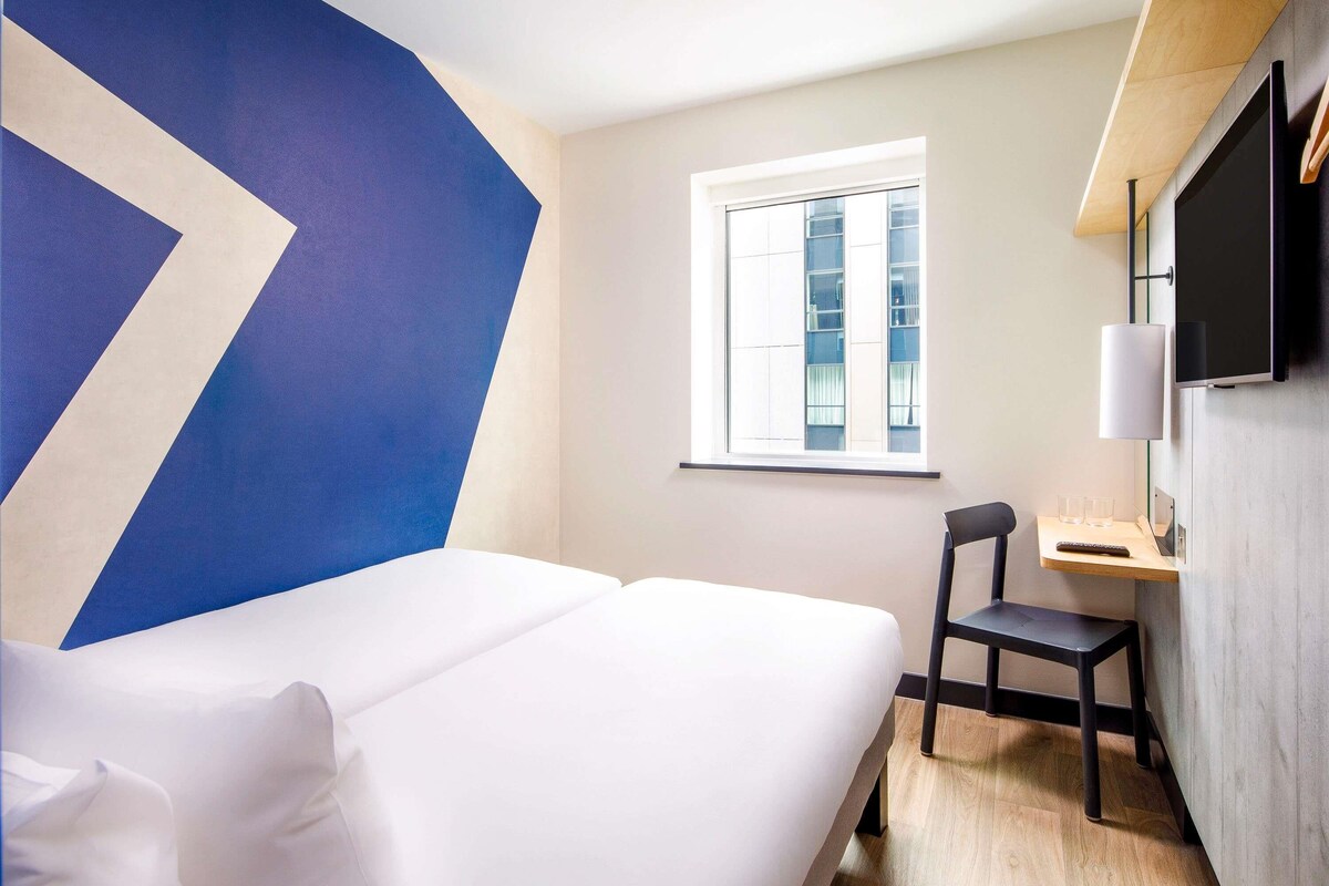 A hotel room with a bed, a desk, a chair, a TV, and a window.