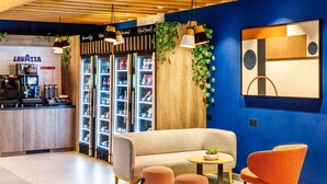 A modern lounge area with a sofa, two ottomans, a coffee machine, and vending machines. - ibis budget Manchester Airport (Manchester)
