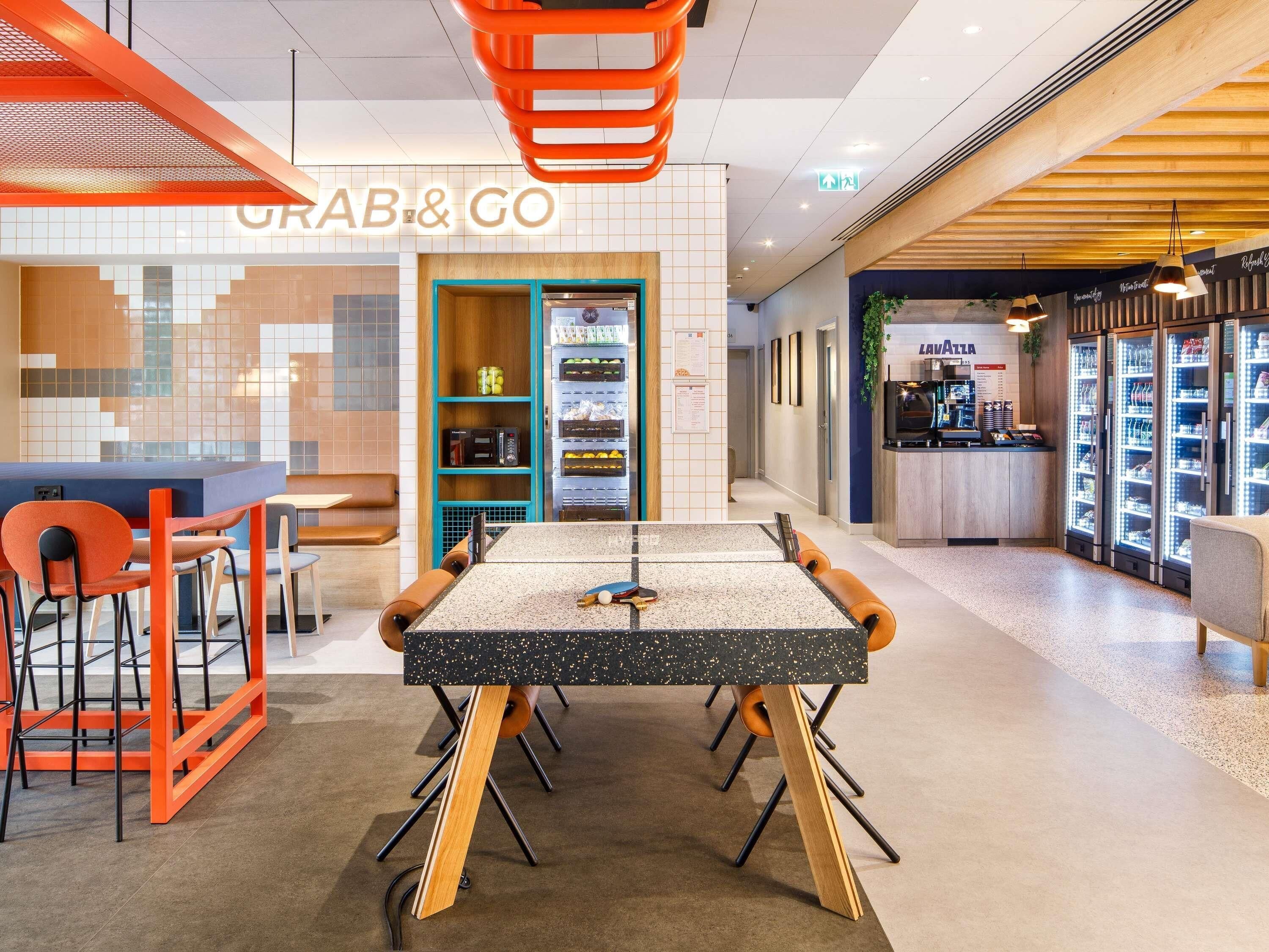 Un interior moderno con una mesa central, sillas naranjas, un mostrador 'Grab & Go' y un puesto de 'Lava Pizza'.