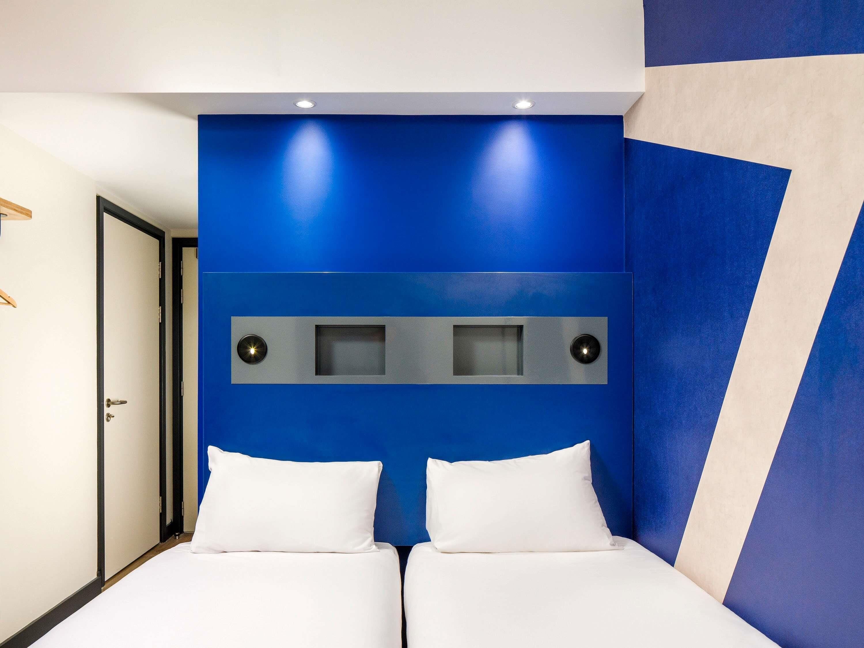 Ein Hotelzimmer mit blauer Akzentwand, einem Bett mit weißer Bettwäsche und einer Tür links.