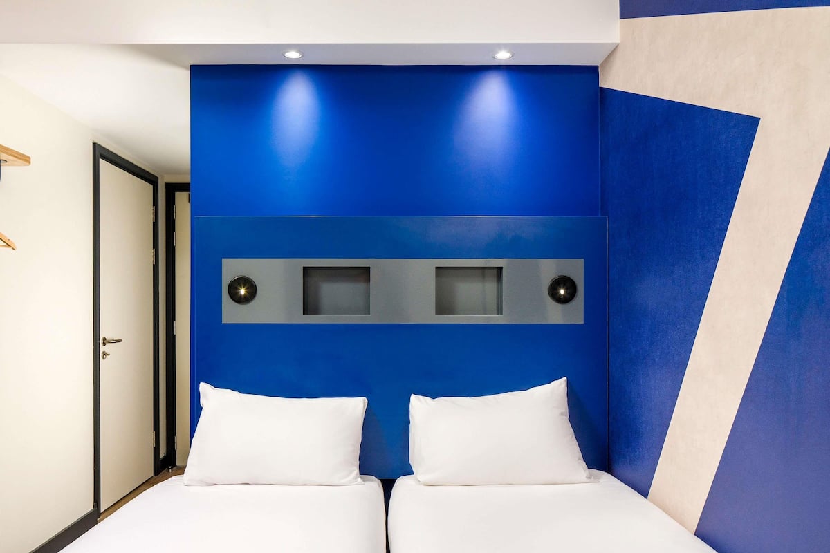 Ein Hotelzimmer mit blauer Akzentwand, einem Bett mit weißer Bettwäsche und einer Tür links.