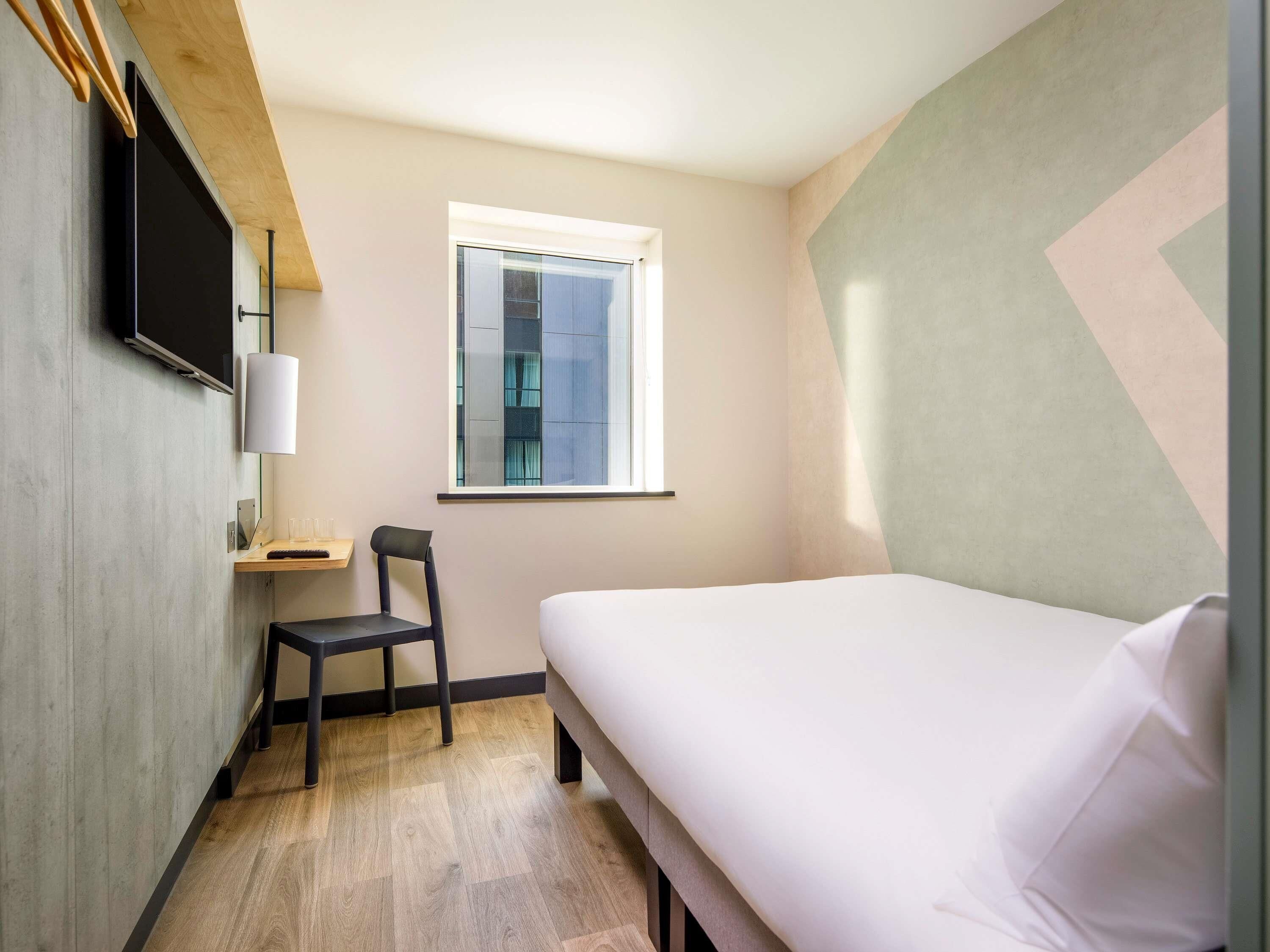 Una camera d'albergo modesta con un letto, una sedia, una TV e una finestra.