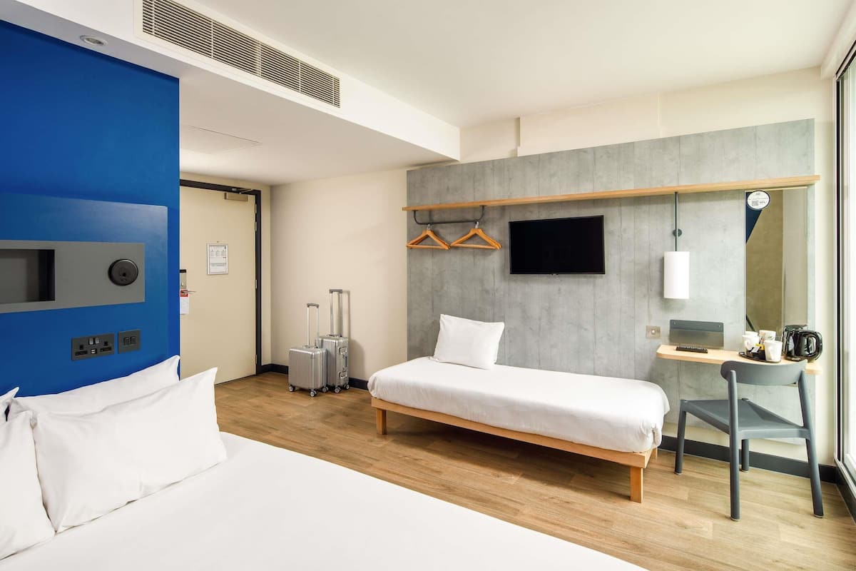 A hotel room with a large bed, a desk with a chair, a TV, and a wardrobe with hangers.