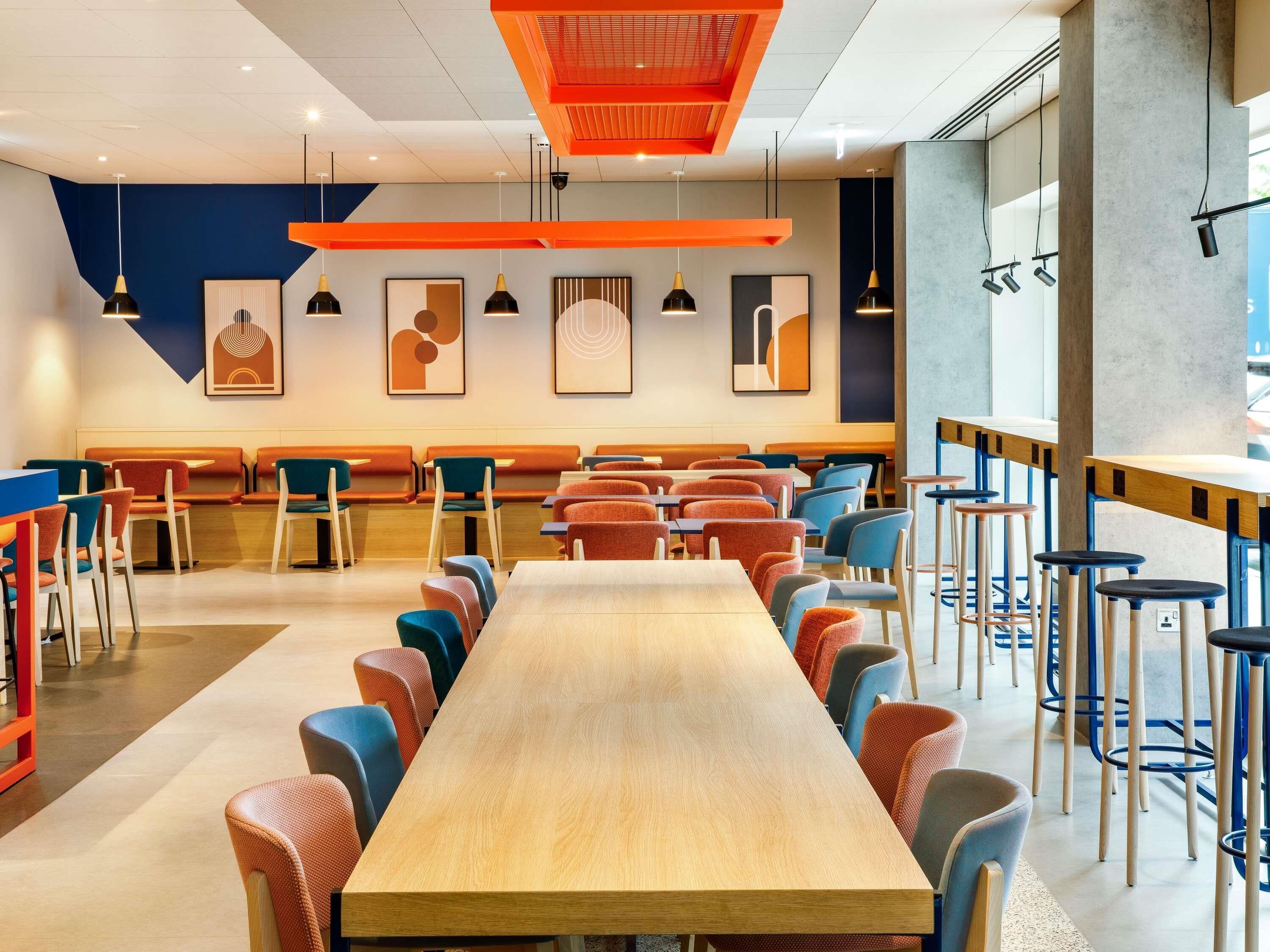 Uma sala de jantar moderna com mesas longas, cadeiras coloridas e iluminação pendente.