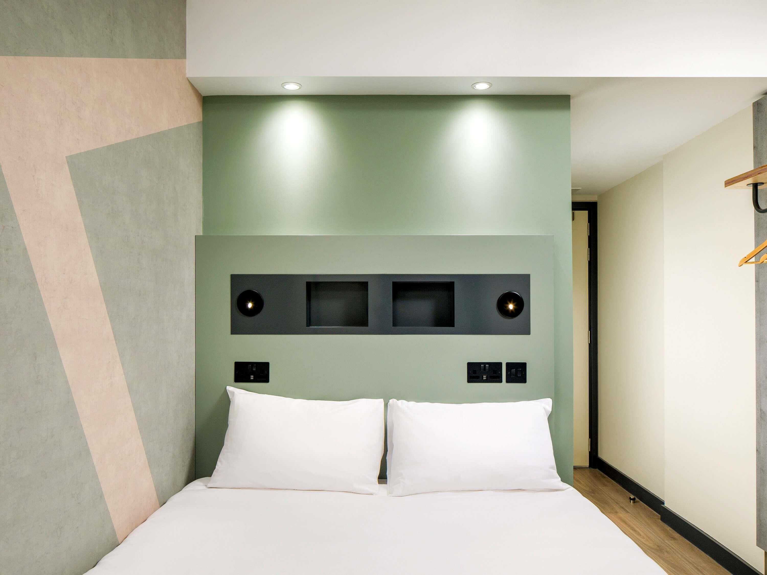 A modern hotel room with a bed, a headboard with built-in lighting, and a wall-mounted control panel.