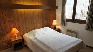 A bedroom with a wooden wall, a bed with a white quilt, two bedside lamps, and a window with curtains. - Chalet, view mountain, tv, 110m², Saint Gervais (Saint-Gervais-les-Bains)