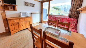 A dining area with a wooden table and chairs, a television on a shelf, and a view of a mountainous landscape through a large window.