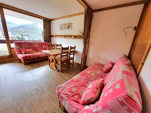 A cozy room with a wooden floor, a red patterned sofa, a wooden dining table with chairs, and a view of mountains through a large window.