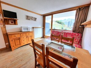 A dining area with a wooden table and chairs, a television on a shelf, and a view of a mountainous landscape through a large window.