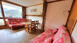 A cozy room with a wooden floor, a red patterned sofa, a wooden dining table with chairs, and a view of mountains through a large window.