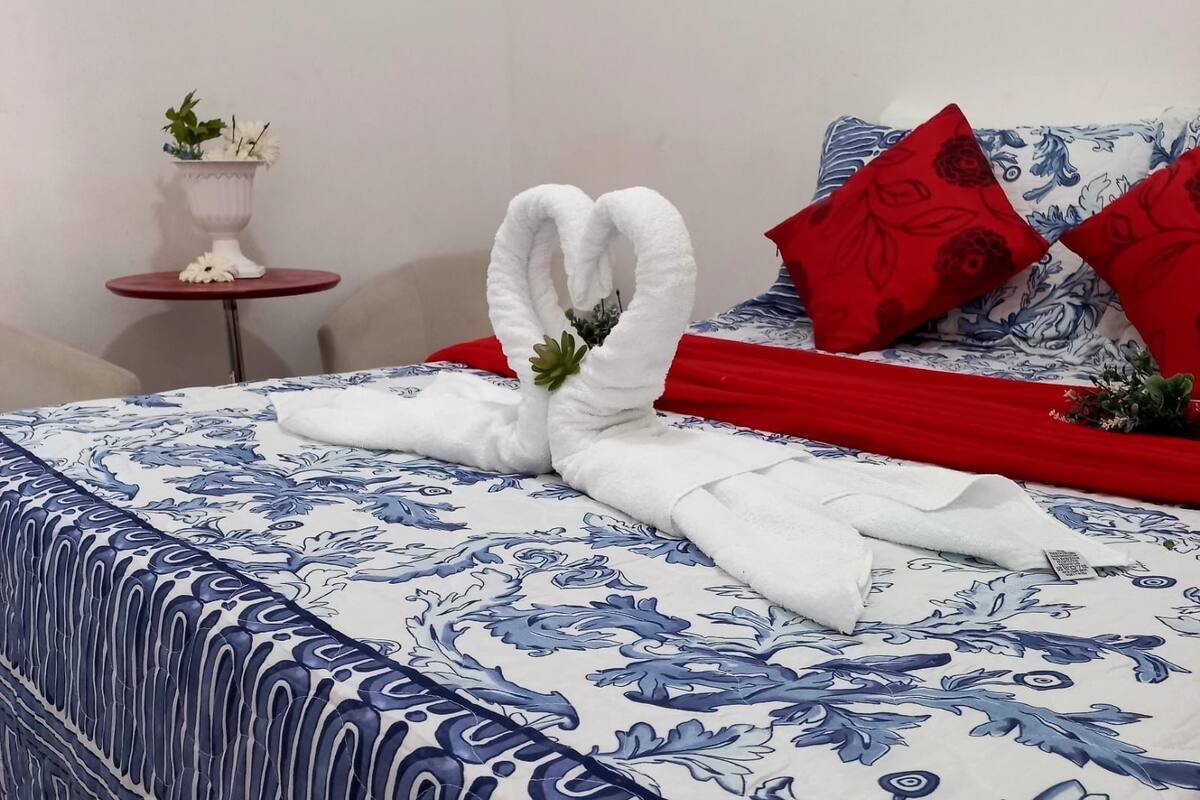 A neatly made bed with a blue and white floral bedspread, red pillows, and a white towel arranged in a swan shape.