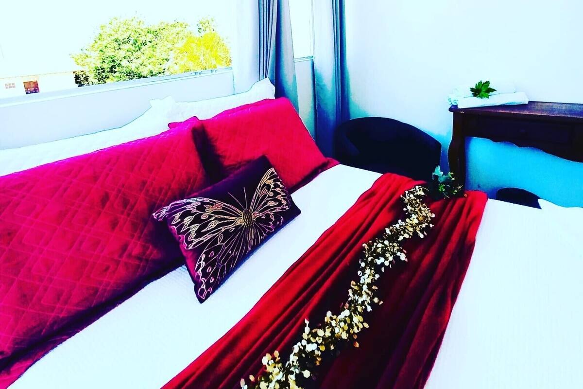 A hotel room with a bed, a window with curtains, a nightstand, and a view of greenery outside.