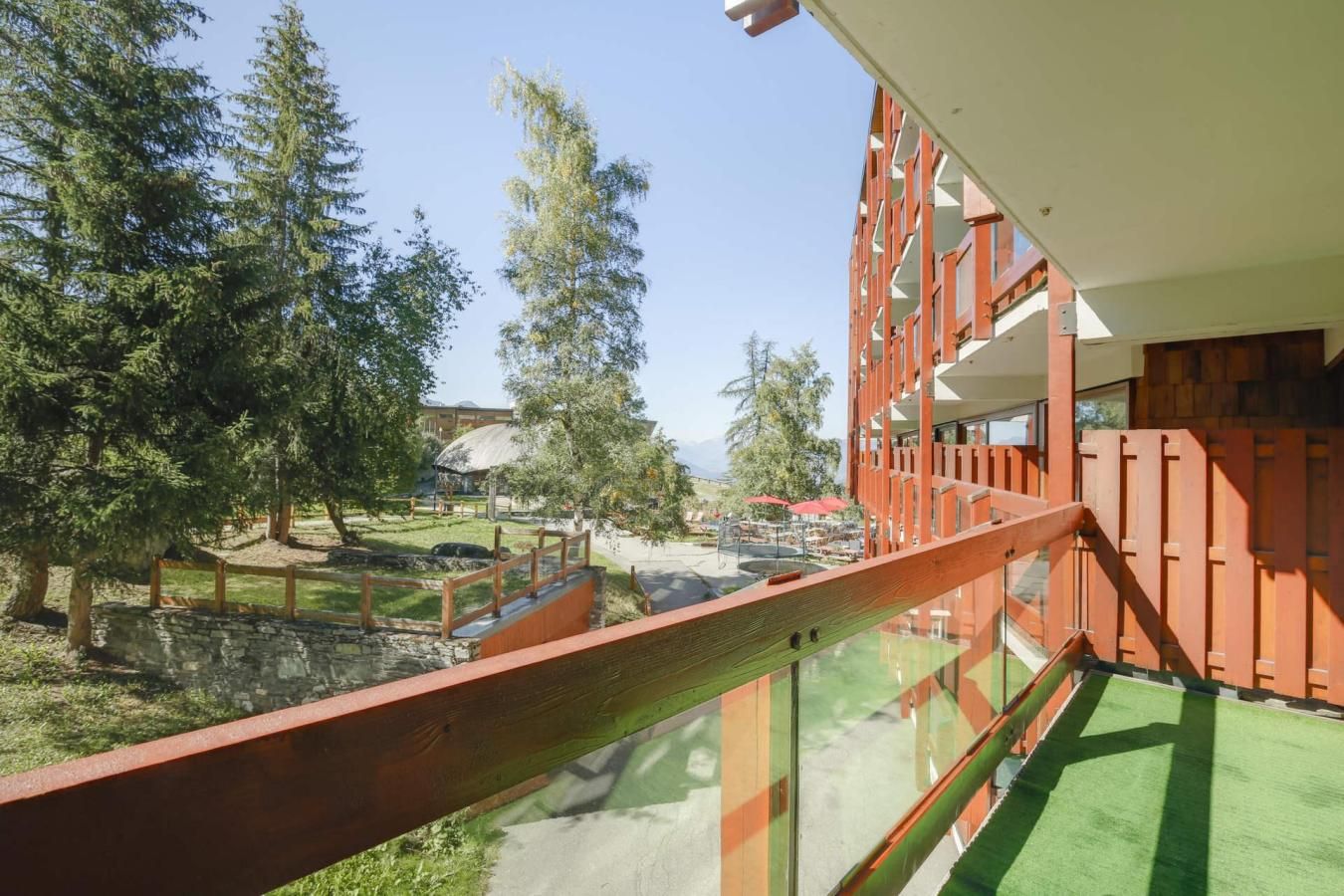 Een modern gebouw met een glazen balkon dat uitkijkt over een aangelegd gebied met bomen en een zwembad.