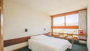 Quarto com uma cama de solteiro, cabeceira de madeira, janela com cortinas e vista para montanhas.