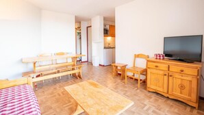 A compact room with a wooden dining table, benches, a TV on a wooden cabinet, and a kitchenette in the background. - 150m from ski slopes, 5th floor, view mountain, balcony, parking, ski locker, 36m², Valfréjus (Valfréjus)