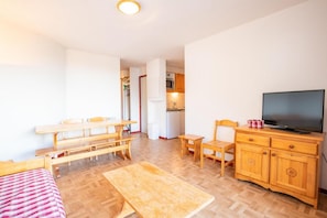 Ein gemütlicher Raum mit Esstisch aus Holz, Bänken, einem Fernseher auf einer Holzkommode und einer kleinen Küche im Hintergrund.