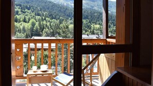 A wooden balcony with a table and chairs, overlooking a forested mountain range.