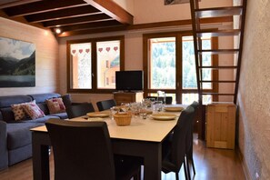 Uma área de refeições acolhedora, com uma mesa de madeira posta para quatro pessoas, um sofá com almofadas e uma televisão sobre um aparador de madeira.