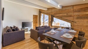 A modern living room with a sofa set, a dining table with chairs, a flat-screen TV, and large windows offering a view of the mountains.