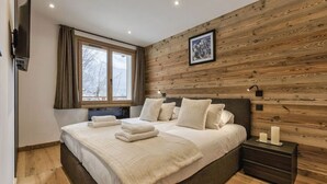 Un dormitorio con pared de madera, una cama grande con ropa blanca, una mesita de noche con velas y una ventana con cortinas.