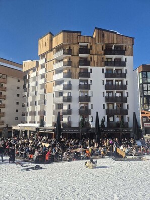 A multi-story building with balconies, a ground-level restaurant, and a snow-covered ground.