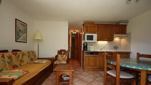A compact living space with a sofa, a small table, a chair, a kitchen area with wooden cabinets and a microwave, and a tiled floor.
