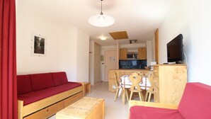 Un espace de vie compact comprenant un canapé en bois, une petite table à manger avec des chaises, un coin cuisine et un téléviseur.