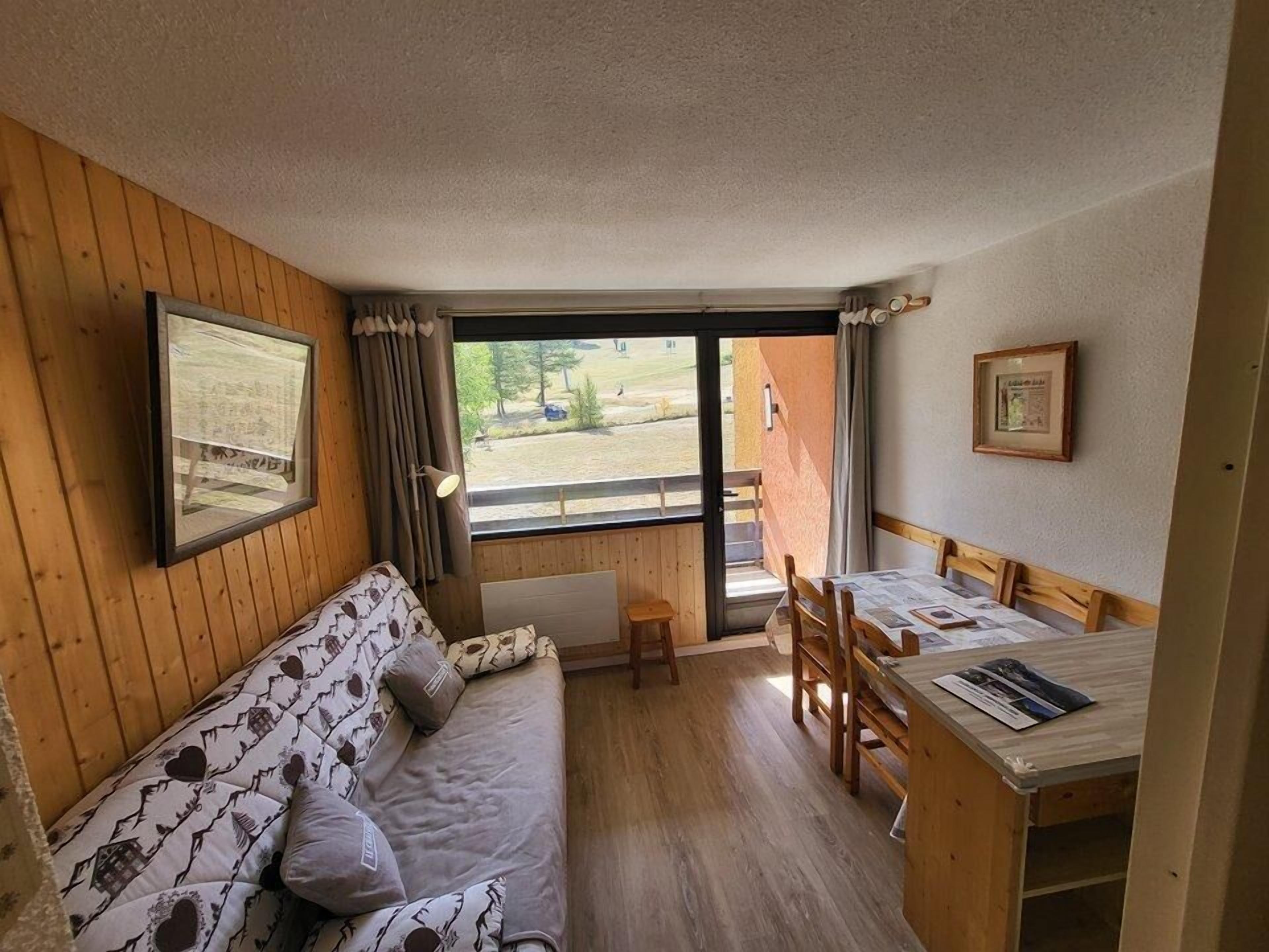 A cozy room with a wooden floor, a sofa with patterned cushions, a table with chairs, and a window offering a view of the outdoors.