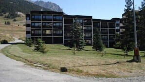 A modern multi-story building with a flat roof, surrounded by a grassy area and a few trees. - 100m from shops, 2nd floor, view mountain, balcony, parking, tv, ski locker, 18m², Montgenèvre (Montgenèvre)