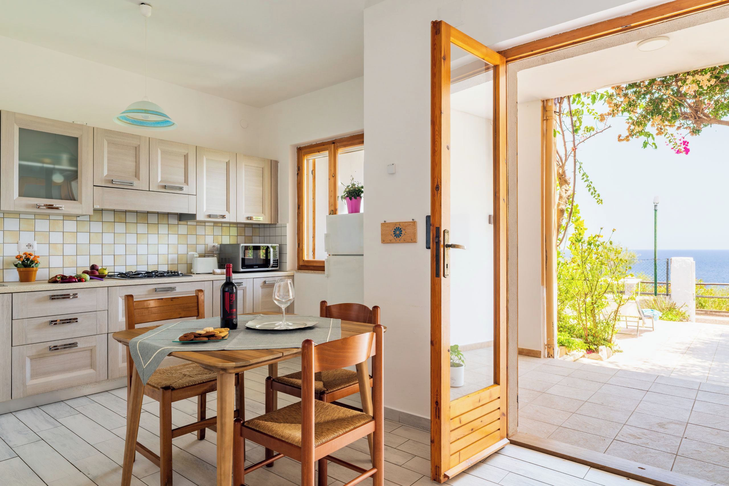 Una cucina con mobili in legno, un tavolo da pranzo con sedie, vista mare tramite una porta scorrevole in vetro e un piccolo balcone con piante.