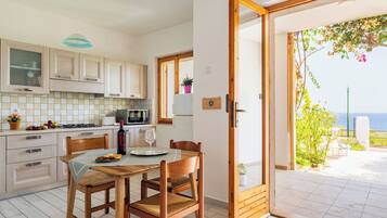 Una cucina con mobili in legno, un tavolo da pranzo con sedie, vista mare tramite una porta scorrevole in vetro e un piccolo balcone con piante.