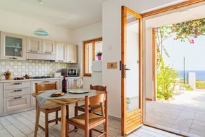 Une cuisine avec des meubles en bois, une table à manger avec des chaises, une vue sur la mer par une porte-fenêtre coulissante et un petit balcon agrémenté de plantes.