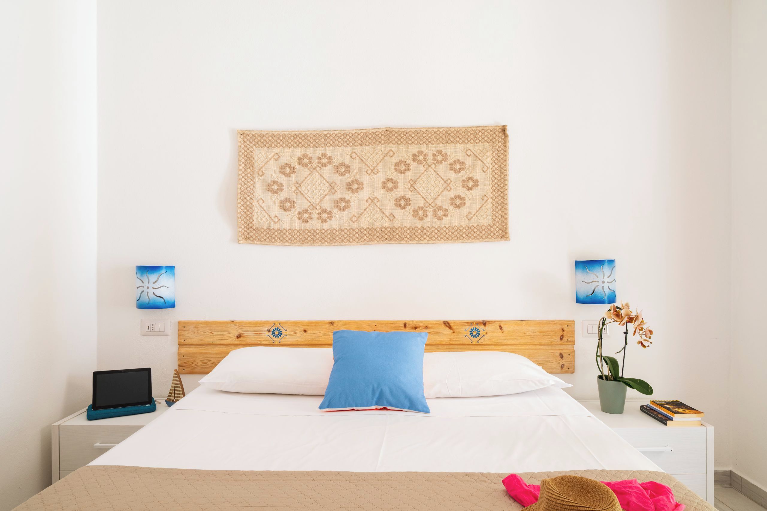 Cama bem arrumada com travesseiro azul, cabeceira de madeira e tapeçaria na parede.