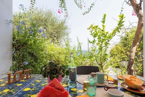 An outdoor dining area with a table set for two, surrounded by lush greenery and a view of the sea.