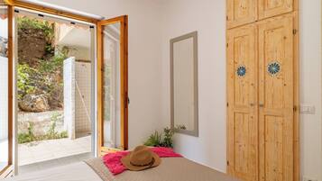 Una habitación con cama, un armario de madera, un espejo y vistas a un espacio exterior con pared de piedra y plantas.