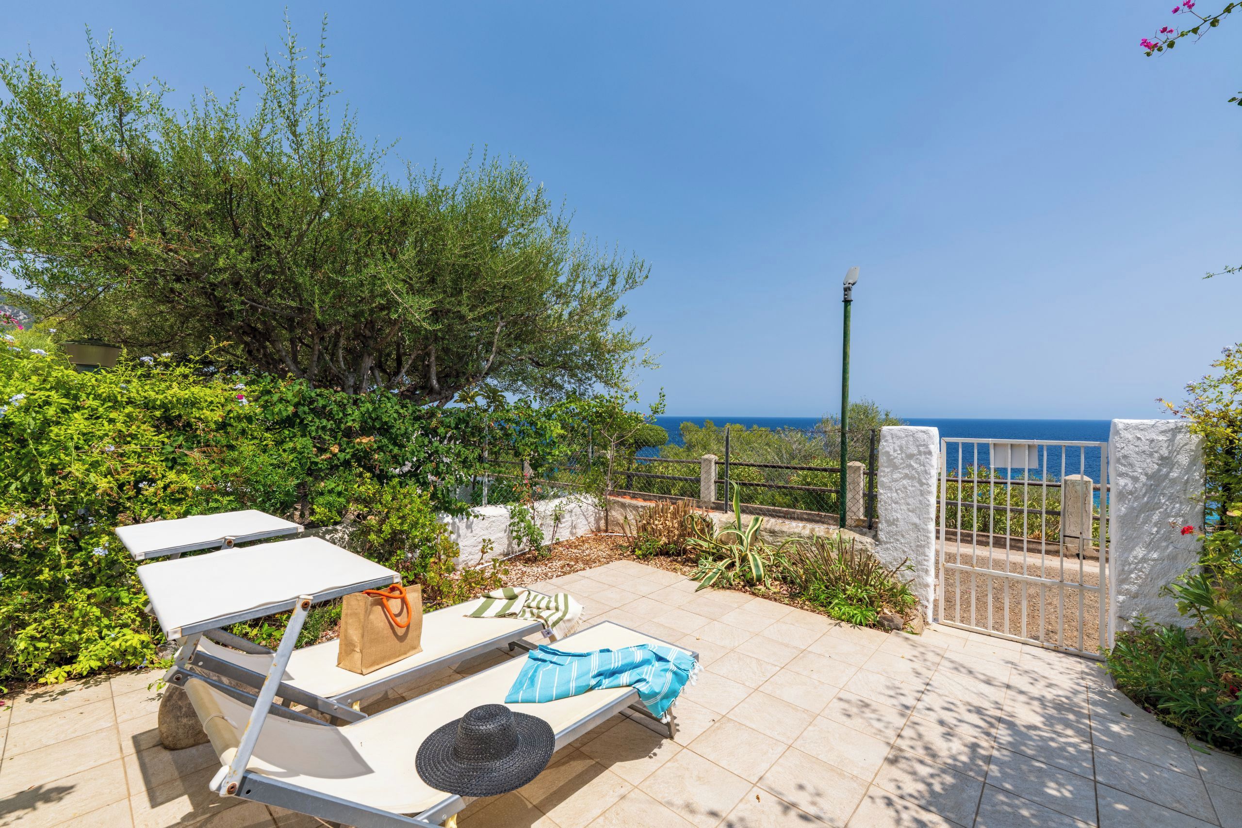A sunlit outdoor seating area with a view of the sea, a metal gate, and a white wall.