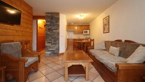 Ein gemütliches Wohnzimmer mit Holzmöbeln, einer Steinsäule, einem Fernseher und einer Küche im Hintergrund.