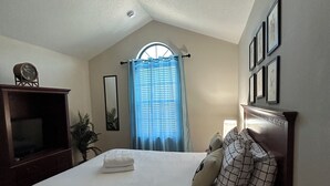 Ein Schlafzimmer mit einem Einzelbett, einem Schminktisch, einer Uhr, einem Fenster mit Vorhängen und gerahmten Bildern an der Wand.