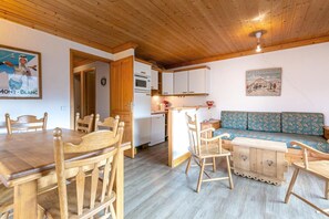 Ein gemütlicher Essbereich mit Holzmöbeln, eine Küche mit weißen Schränken und ein blaues Sofa mit Muster.