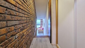 Un pasillo estrecho con una pared de ladrillo a un lado y una pared blanca al otro, que conduce a una habitación con una cama y un ventanal.