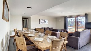 A dining area with a long wooden table set for six, surrounded by cushioned chairs. A large window provides a view of the outdoors. - 200m from ski slopes, Ground floor, balcony, tv, 90m², Les Menuires (Les Menuires)