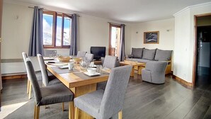 Moderni olohuone, jossa on puinen ruokapöytä, harmaalla kankaalla verhoillut tuolit, sohva ja televisio.
