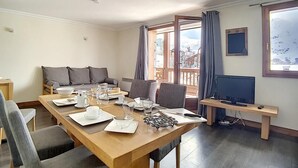 Un salon moderne avec une table à manger en bois, préparée pour quatre personnes, un canapé avec des coussins, un téléviseur posé sur un meuble et un balcon offrant une vue sur les montagnes.