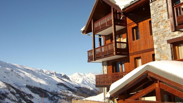 Un edificio de madera de varios pisos con balcones, paredes de piedra y una montaña nevada al fondo.