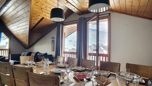 Un comedor con mesa de madera preparada para seis personas, ventanas amplias con cortinas y vistas a montañas nevadas.