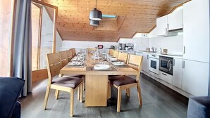 Una cocina moderna con armarios blancos y electrodomésticos de acero inoxidable, un área de comedor con una mesa de madera preparada para seis comensales, y un techo inclinado con vigas de madera a la vista.