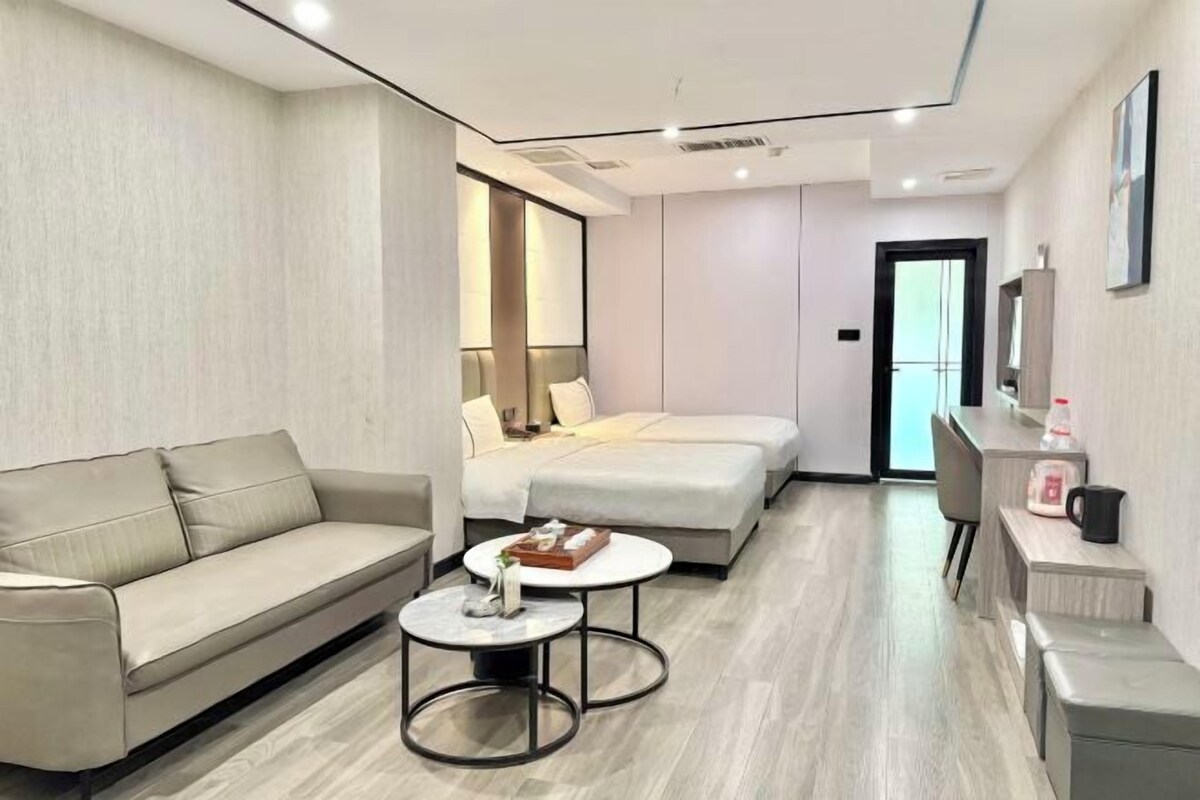 A modern hotel room with a sofa, two beds, a small table, and a desk with a chair.