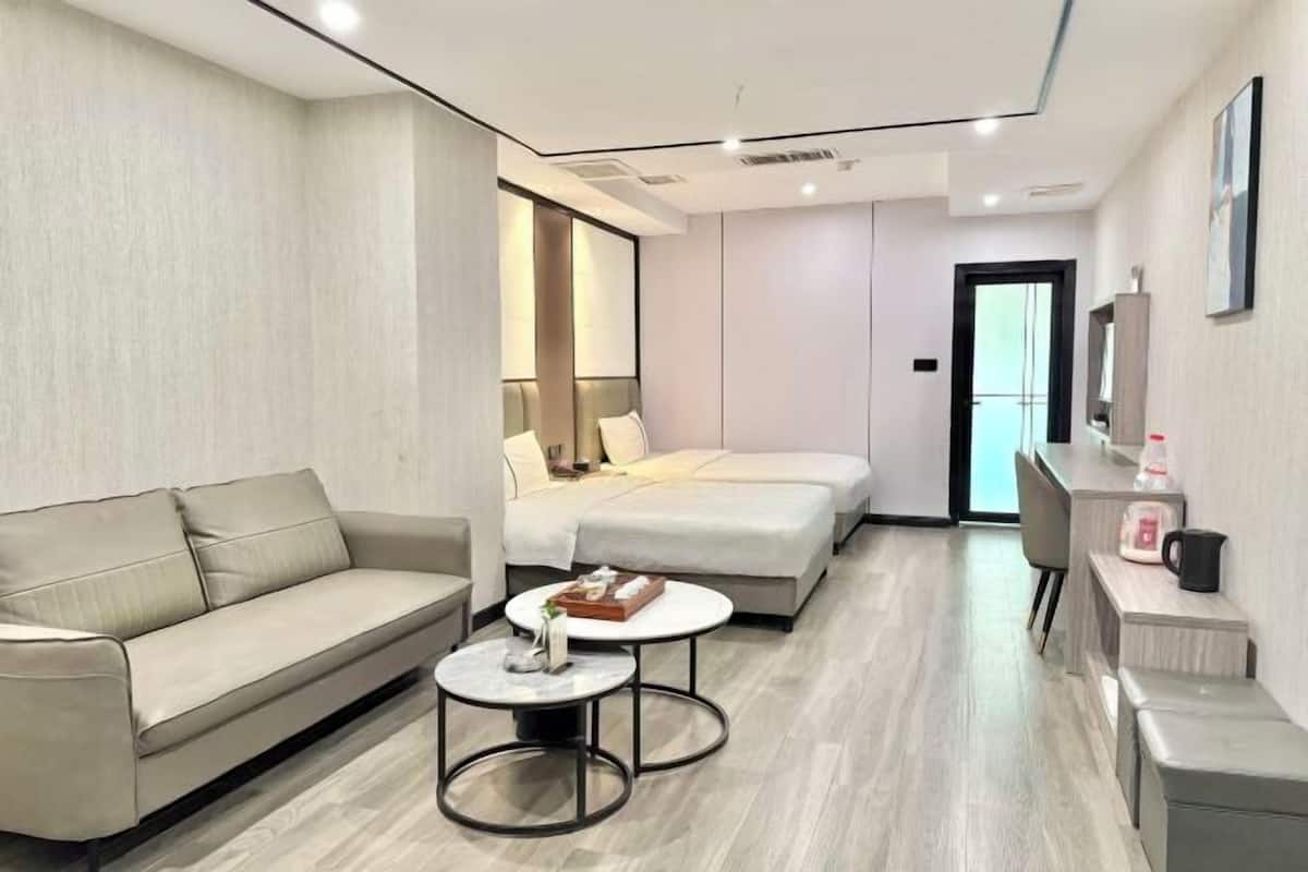 A modern hotel room with a sofa, two beds, a small table, and a desk with a chair.