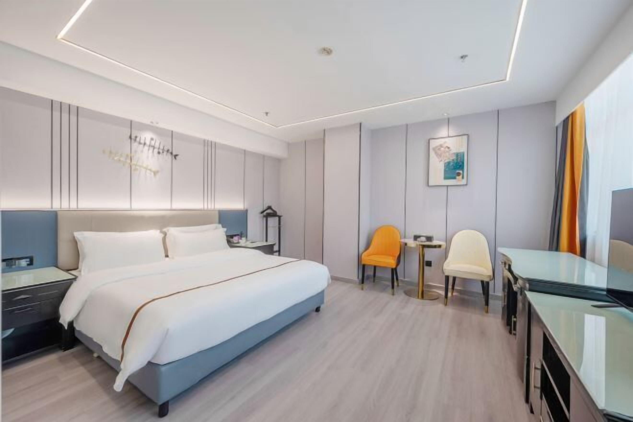 A modern hotel room with a large bed, a desk, two chairs, a small table, and a wall-mounted artwork.